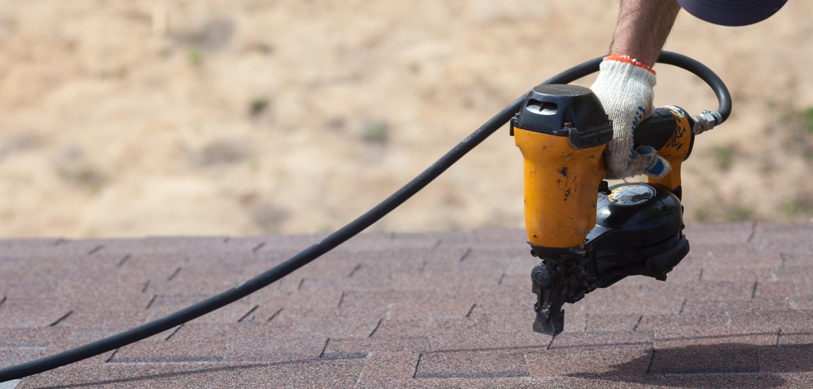 Roofer builder worker with nailgun installing Asphalt Shingles or Bitumen Tiles on a new house under construction